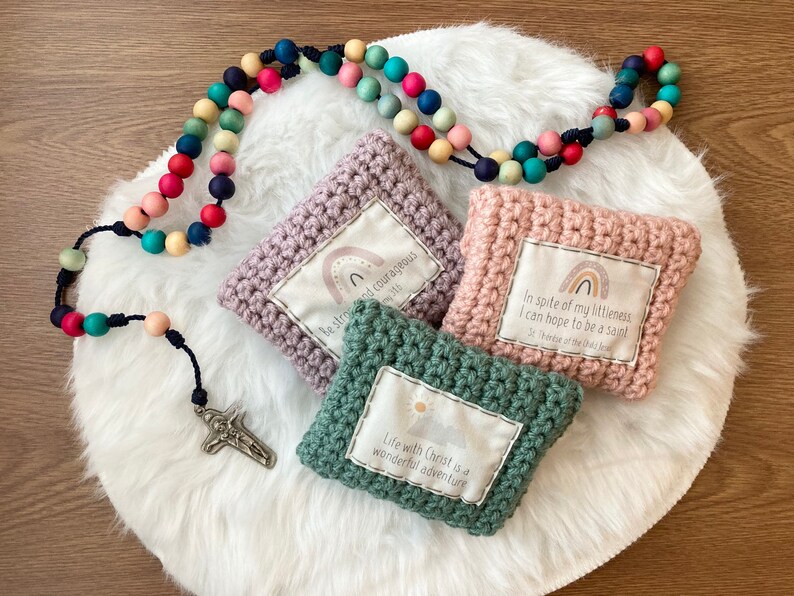 Peut inclure: Trois pochettes en crochet avec des citations inspirantes et des motifs arc-en-ciel. Les pochettes sont rose, violette et verte. Un chapelet en bois color&eacute; avec une croix en argent est sur une surface blanche et moelleuse.