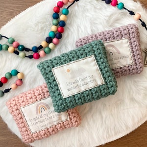 Peut inclure: Trois pochettes en crochet avec des citations inspirantes et des motifs arc-en-ciel. Les pochettes sont vertes, roses et violettes. Les citations sont "La vie avec le Christ est une merveilleuse aventure", "Malgr&eacute; ma petite taille, je peux esp&eacute;rer &ecirc;tre un saint", et "Soyez gentil et courageux".
