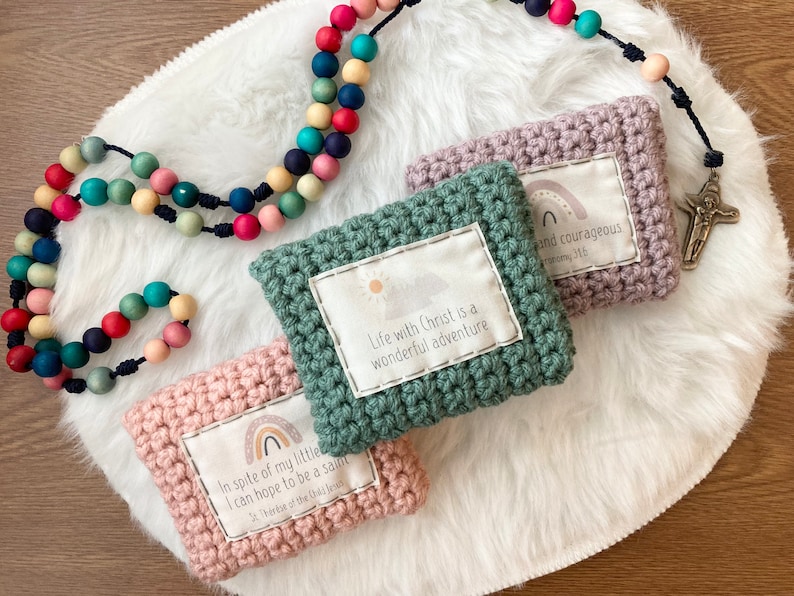 Peut inclure: Trois pochettes en crochet avec des citations inspirantes et des motifs arc-en-ciel. Les pochettes sont vertes, roses et violettes. Les citations sont "La vie avec le Christ est une merveilleuse aventure", "Malgr&eacute; ma petite taille, je peux esp&eacute;rer &ecirc;tre un saint", et "Soyez gentil et courageux".