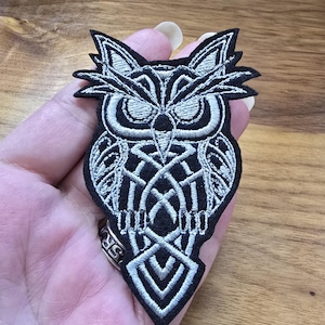 May include: An embroidered patch featuring a stylised owl design. The owl is rendered in silver-coloured thread against a solid black fabric background, showcasing intricate geometric patterns and sharp, defined lines throughout the entire bird figure.