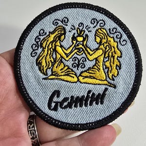 May include: A circular embroidered patch featuring a light blue denim-like background with a black stitched border. The design depicts two yellow figures with long hair facing each other, holding a small yellow object. The word Gemini is stitched in black.
