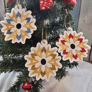 May include: Three white snowflake decorations with gold, blue, and red detailing hang on a Christmas tree. Each ornament has a black centre and is trimmed with white stitching. The decorations are suspended by twine.