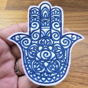 May include: A Hamsa hand patch featuring intricate dark blue embroidery on a white felt background. The design includes detailed swirling patterns, geometric shapes, and a central eye motif, all outlined in a thick white border.