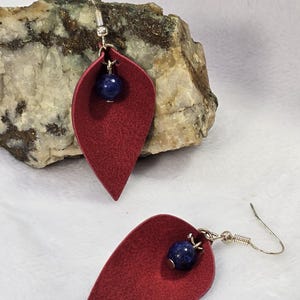 May include: A pair of red leaf-shaped earrings with a dark blue bead accent. The earrings have silver-colored ear wires. The earrings are displayed on a rock with a white background.