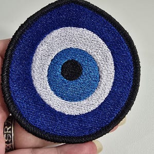 May include: A circular embroidered patch featuring a stylized eye design. The center is a solid black circle, surrounded by a medium blue ring, a wider white ring, and an outer dark blue ring, all enclosed by a thick black stitched border.