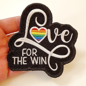 May include: A black embroidered patch featuring the white cursive text Love with a rainbow-coloured heart replacing the letter o, and the white block text FOR THE WIN underneath, all set against a black background with a stitched border.