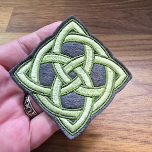 May include: A diamond-shaped embroidered patch featuring a lime green Celtic knot design stitched onto a dark gray felt base. The intricate, interlacing pattern creates a continuous loop effect, with the green thread contrasting against the dark gray fabric background.