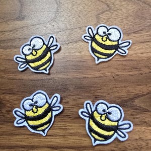 May include: Four embroidered bee patches featuring a cartoon design with large white eyes, yellow and black striped bodies, and white wings. Each patch has a white border and is arranged in a grid pattern on a brown wood grain surface.