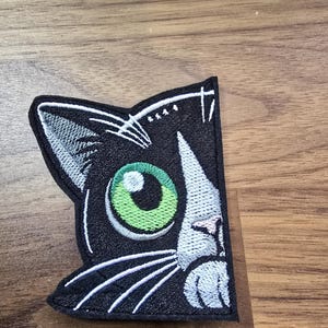 May include: An embroidered patch featuring a black cat head with a large, bright green eye, white whiskers, and a white patch on the nose and forehead. The design is outlined in black thread with a clean, finished edge on a dark fabric base.