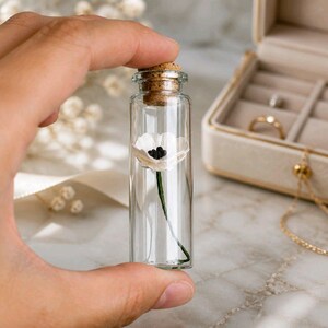 May include: A clear glass cylindrical vial with a cork stopper containing a single miniature white flower with a dark center and a thin green stem. The vial is transparent, showcasing the delicate floral detail preserved inside the glass container.