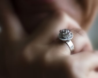 May include: A silver ring with a small teacup and saucer design. The ring is worn on a person's finger.