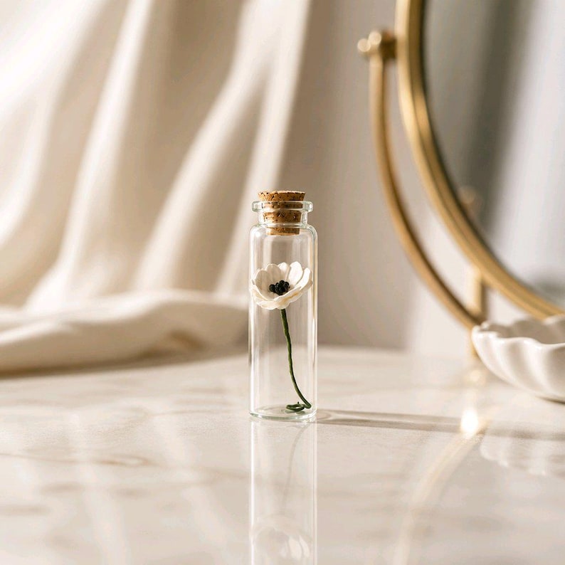 May include: A clear glass vial with a cork stopper containing a single small white flower with a dark center and a thin green stem. The miniature floral arrangement is encased within the transparent cylindrical bottle, standing upright.