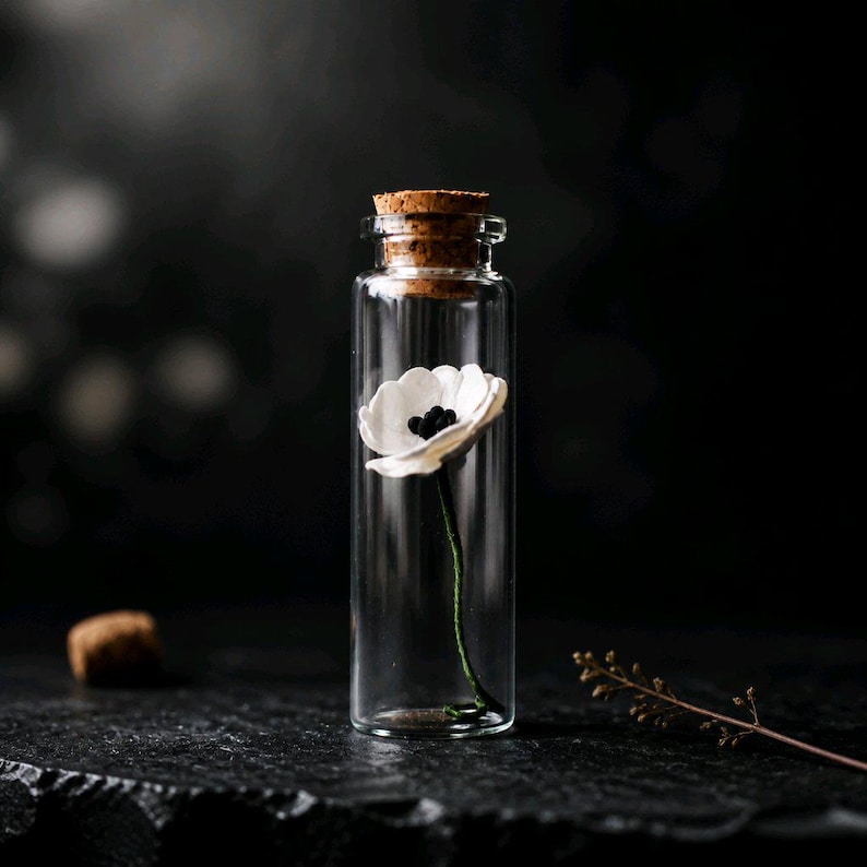 May include: A clear glass vial with a cork stopper containing a single white flower with a dark center and a thin green stem. The flower is positioned upright inside the transparent cylindrical container, showcasing a delicate and simple botanical design.