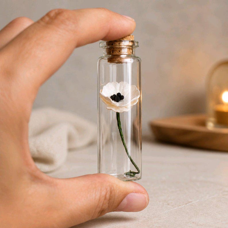 May include: A small, clear glass vial sealed with a cork stopper, containing a single miniature white flower with a dark center and a thin green stem. The transparent cylindrical bottle is held upright, showcasing the delicate floral detail inside.
