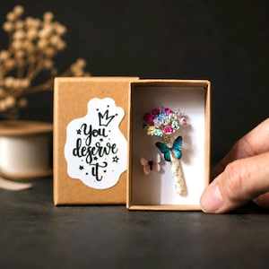 Może przedstawiać: Małe, otwarte kartonowe pudełko z miniaturowym bukietem kolorowych kwiat&oacute;w i dwoma motylami. Pudełko ma naklejkę z napisem "You deserve it". Motyle są niebieskie i r&oacute;żowe. Tło jest ciemne.