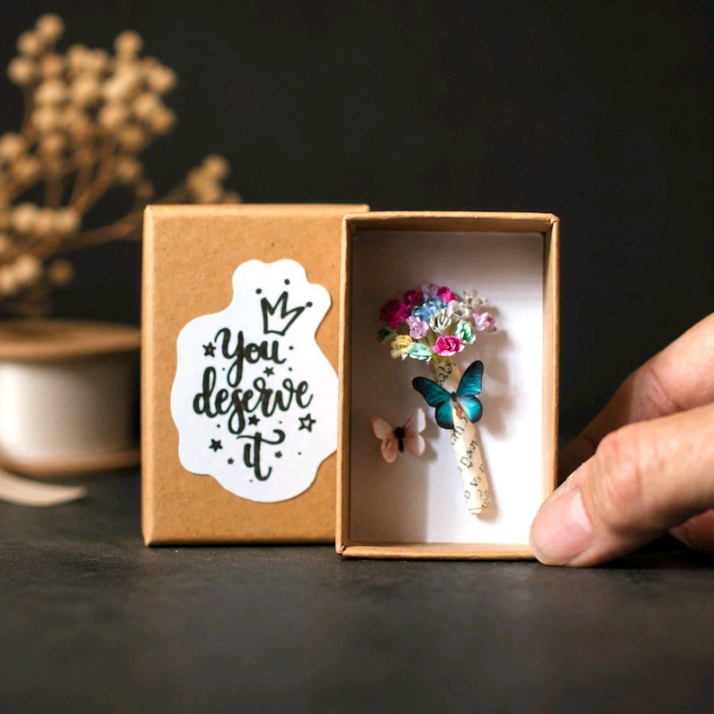Może przedstawiać: Małe, otwarte kartonowe pudełko z miniaturowym bukietem kolorowych kwiat&oacute;w i dwoma motylami. Pudełko ma naklejkę z napisem "You deserve it". Motyle są niebieskie i r&oacute;żowe. Tło jest ciemne.