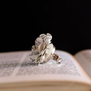 Peut inclure: Une bague en argent avec une fleur en papier blanc faite de pages de livre. La bague repose sur un livre ouvert.