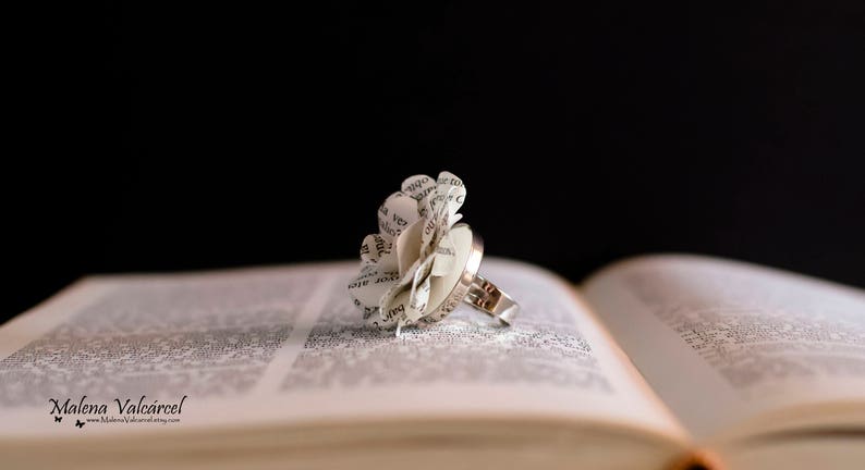 Peut inclure: Une bague en argent avec une fleur en papier blanc faite de pages de livre. La bague repose sur un livre ouvert.