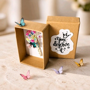 Może przedstawiać: Brązowe pudełko na prezent z białą naklejką z napisem "You deserve it". Wewnątrz wystawiony jest miniaturowy bukiet z kolorowych papierowych kwiat&oacute;w i motyli. Kilka małych dekoracji motyli jest rozrzuconych wok&oacute;ł pudełka.