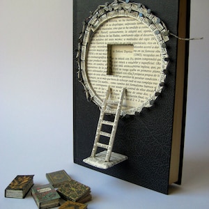 May include: A black book with a paper ladder attached to the front cover. The ladder is made of pages from a book and is decorated with small metal tacks. The book is surrounded by small, colorful, miniature books. The text on the book cover reads "There is no frigate like a book..."