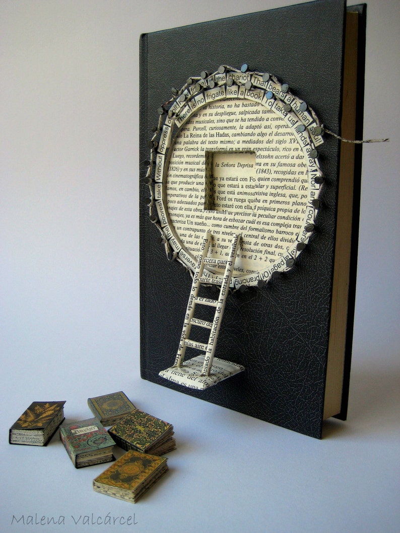 May include: A black book with a paper ladder attached to the front cover. The ladder is made of pages from a book and is decorated with small metal tacks. The book is surrounded by small, colorful, miniature books. The text on the book cover reads "There is no frigate like a book..."