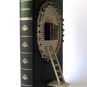 May include: A black book with a gold spine and a paper wheel attached to the front cover. The wheel is made of pages from a book and has a ladder attached to it. The book's spine has the text "Shakespeare Comedias" and "RBA Editores 4".