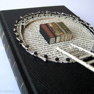 May include: A black book with a paper ladder and a circle of paper scraps around it. The paper scraps are attached to the book with black tacks. The paper scraps have text on them, including "Without Oppress of" and "Take us sandal away".