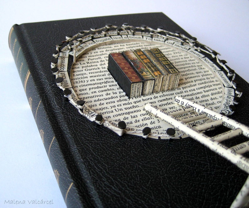 May include: A black book with a paper ladder and a circle of paper scraps around it. The paper scraps are attached to the book with black tacks. The paper scraps have text on them, including "Without Oppress of" and "Take us sandal away".