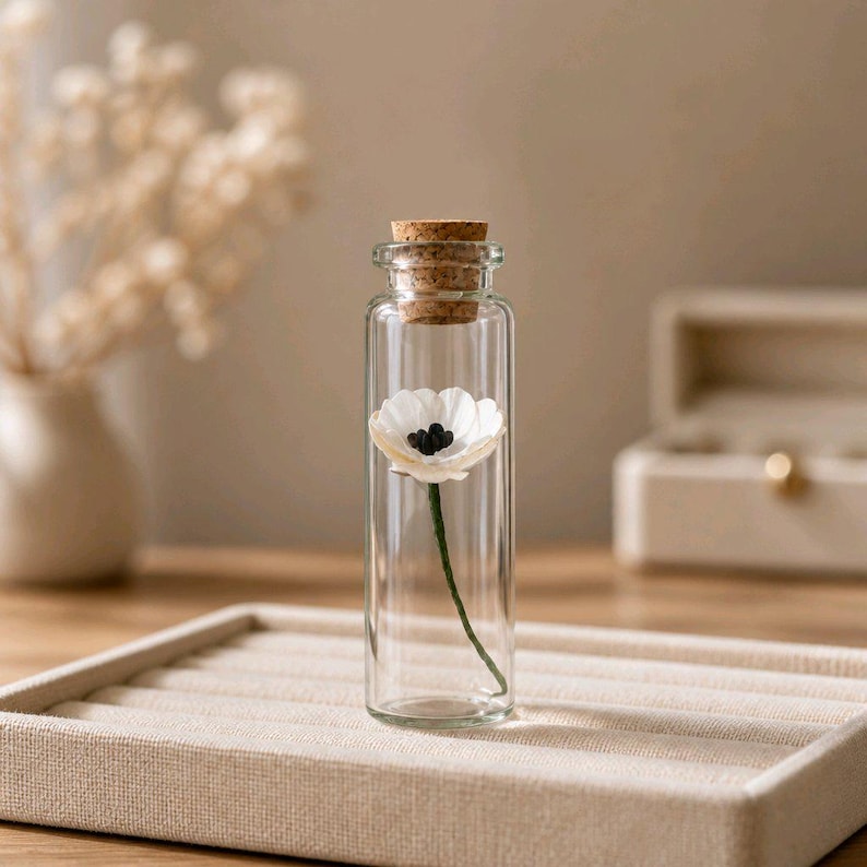 May include: A clear glass cylindrical vial with a cork stopper containing a single white flower with a dark center and a thin green stem. The glass bottle is transparent, showcasing the delicate floral specimen inside the narrow vessel.