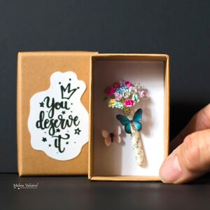 Może przedstawiać: Małe, otwarte pudełko z miniaturowym bukietem kolorowych papierowych kwiat&oacute;w i dwiema dekoracjami motyli. Pudełko ma naklejkę z napisem "You deserve it". Tło jest ciemne.