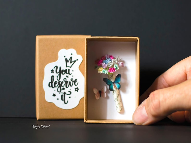 Może przedstawiać: Małe, otwarte pudełko z miniaturowym bukietem kolorowych papierowych kwiat&oacute;w i dwiema dekoracjami motyli. Pudełko ma naklejkę z napisem "You deserve it". Tło jest ciemne.
