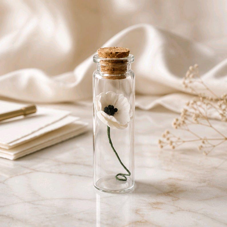 May include: A clear glass cylindrical vial sealed with a light brown cork stopper. Inside the vial is a single white flower with a dark center and a thin green stem that curls at the bottom, resting against the interior base of the glass container.