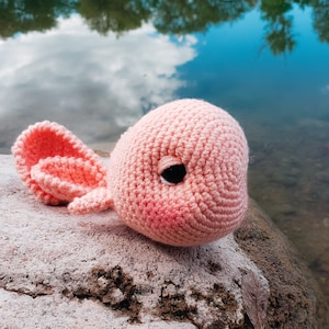 May include: A pink crocheted axolotl plush toy with a black eye. The toy is sitting on a rock near a body of water.
