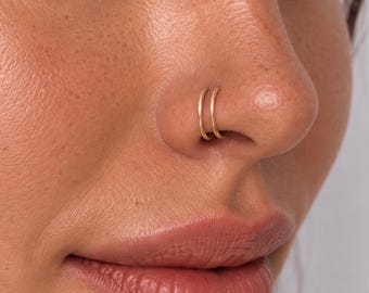 May include: A double hoop nose ring made of thin, polished gold-toned metal. The jewelry consists of two separate, circular bands that sit side-by-side, creating the appearance of a stacked or layered look when worn in the nostril piercing.
