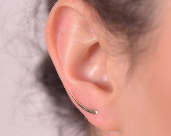 May include: A close-up of a person's ear with a gold-colored ear climber earring. The earring curves upward along the ear, with a simple, minimalist design. The earring is a delicate piece of jewelry.