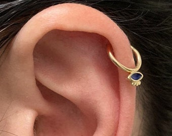 May include: Gold hoop earring with a small blue evil eye charm. The earring is worn in the upper cartilage of the ear.