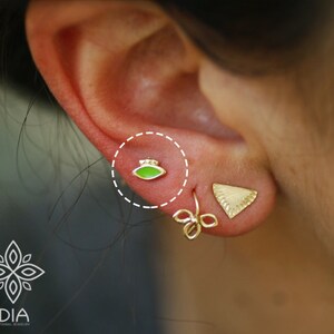 May include: A close-up of a person's ear with two earrings. The earring in the lower lobe is a gold triangle shape. The earring in the upper lobe is a small green oval.