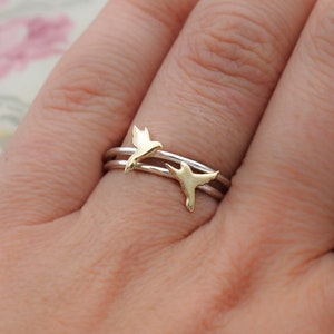 May include: A silver ring with two gold hummingbird charms. The ring is worn on a person's finger.