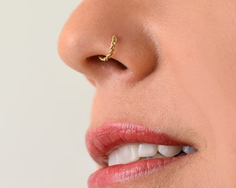 May include: A close-up of a person's face with a gold nose ring. The ring is a braided design. The person is smiling and has their mouth slightly open.