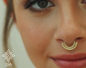 May include: A close-up of a person's face with a gold septum ring. The ring is a half-circle shape with a beaded edge.