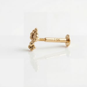 Pode incluir: Um brinco labret dourado 14K. O brinco apresenta um aglomerado de pequenas contas redondas em uma extremidade e um disco plano na outra. A haste tem um design roscado. O texto "14K" é visível na haste.