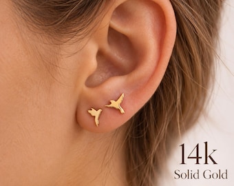 May include: A pair of 14k solid gold stud earrings shaped like small flying birds. The earrings feature a polished, reflective finish and are worn on the earlobe. The text 14k Solid Gold is visible in the bottom right corner of the image.