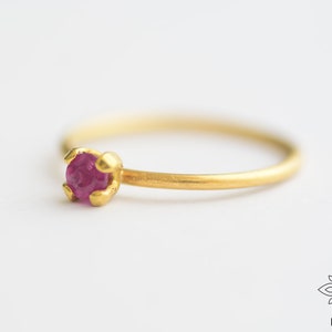 May include: A delicate gold ring with a small, round, pink ruby gemstone set in a simple prong setting.