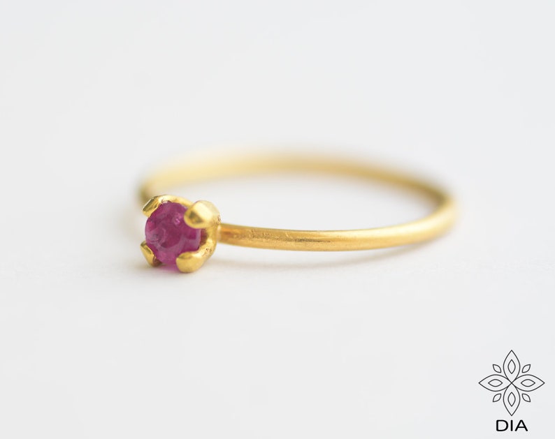 May include: A delicate gold ring with a small, round, pink ruby gemstone set in a simple prong setting.