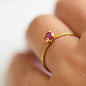 May include: A delicate gold ring with a small, round, pink gemstone set in a simple, claw setting.