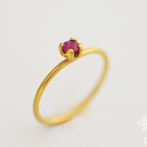 May include: A delicate gold ring with a single, small, ruby-red gemstone set in a prong setting.