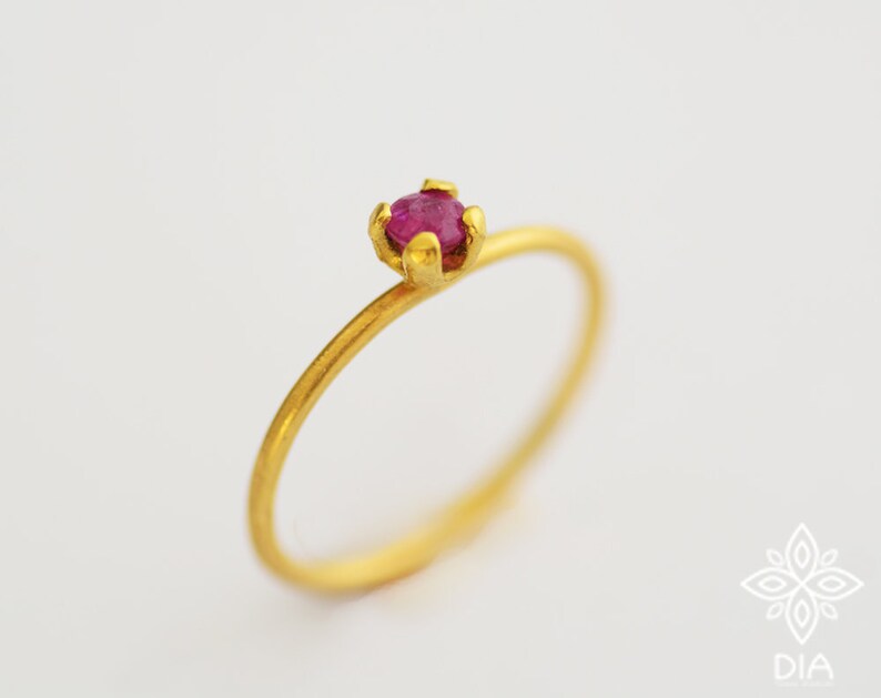 May include: A delicate gold ring with a single, small, ruby-red gemstone set in a prong setting.