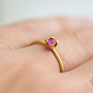May include: A delicate gold ring with a single, small, round, pink gemstone set in a prong setting.