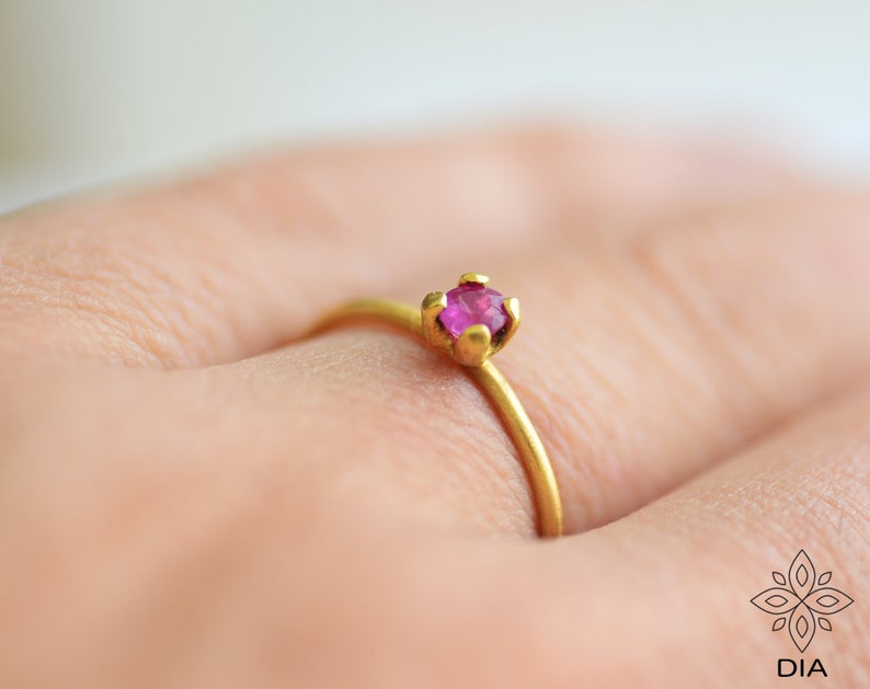 May include: A delicate gold ring with a single, small, round, pink gemstone set in a prong setting.