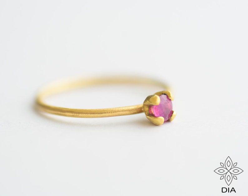 May include: A delicate gold ring with a single, small, pink gemstone set in a claw setting.